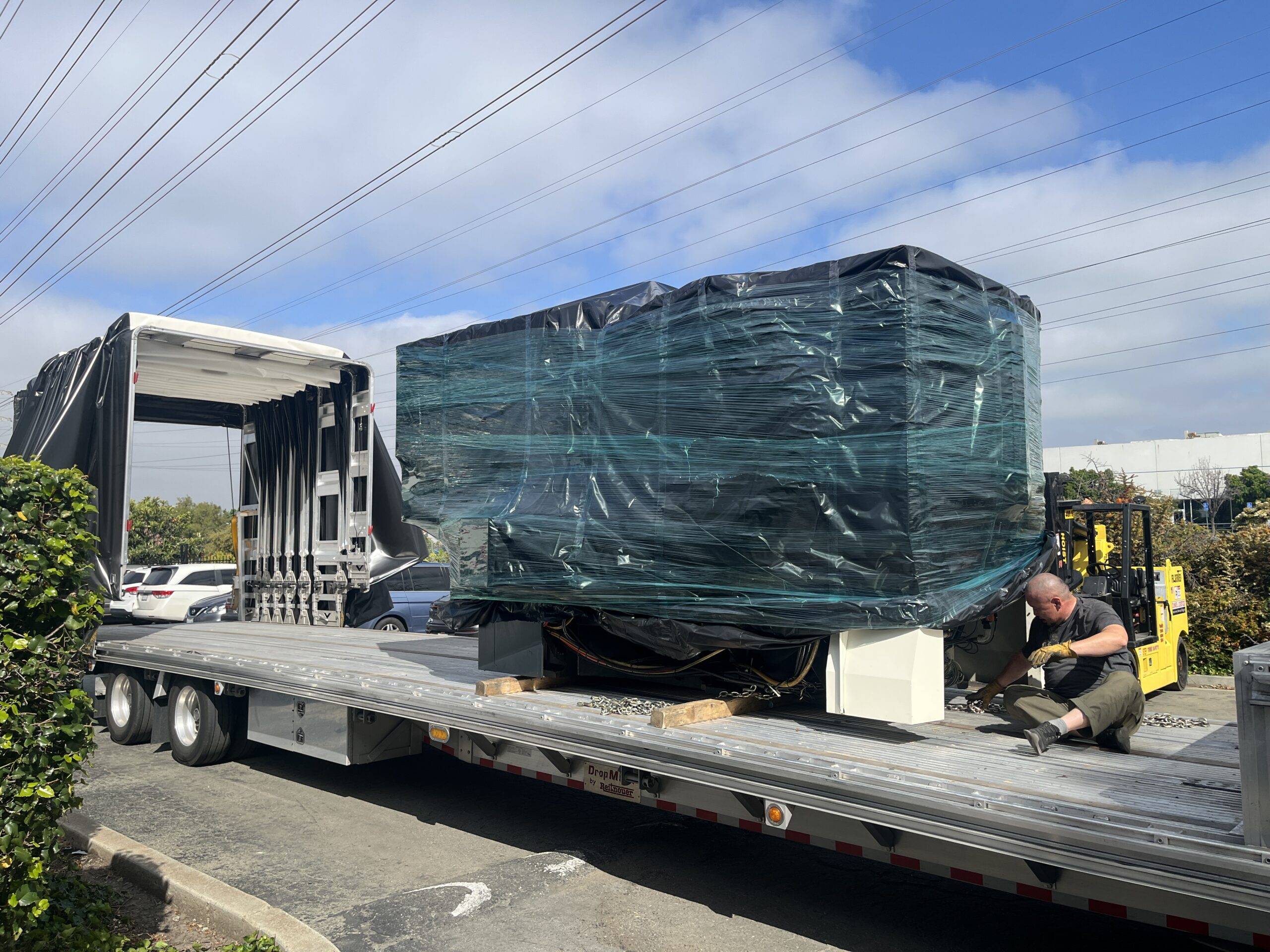 Rigger unloading a large machine from a step deck conestoga trailer using a forklift