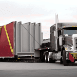 Double drop heavy haul trailer transporting a large industrial generator on a highway.