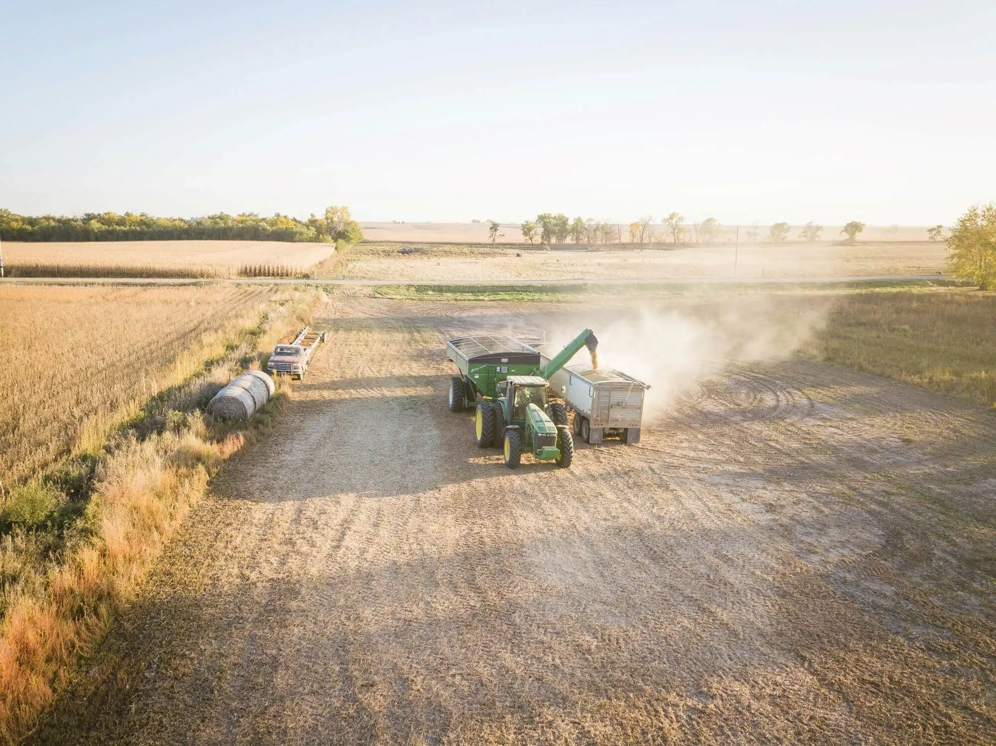 Green tractor unloading harvested grain into a bulk hopper trailer in a sunlit field for agricultural freight transport