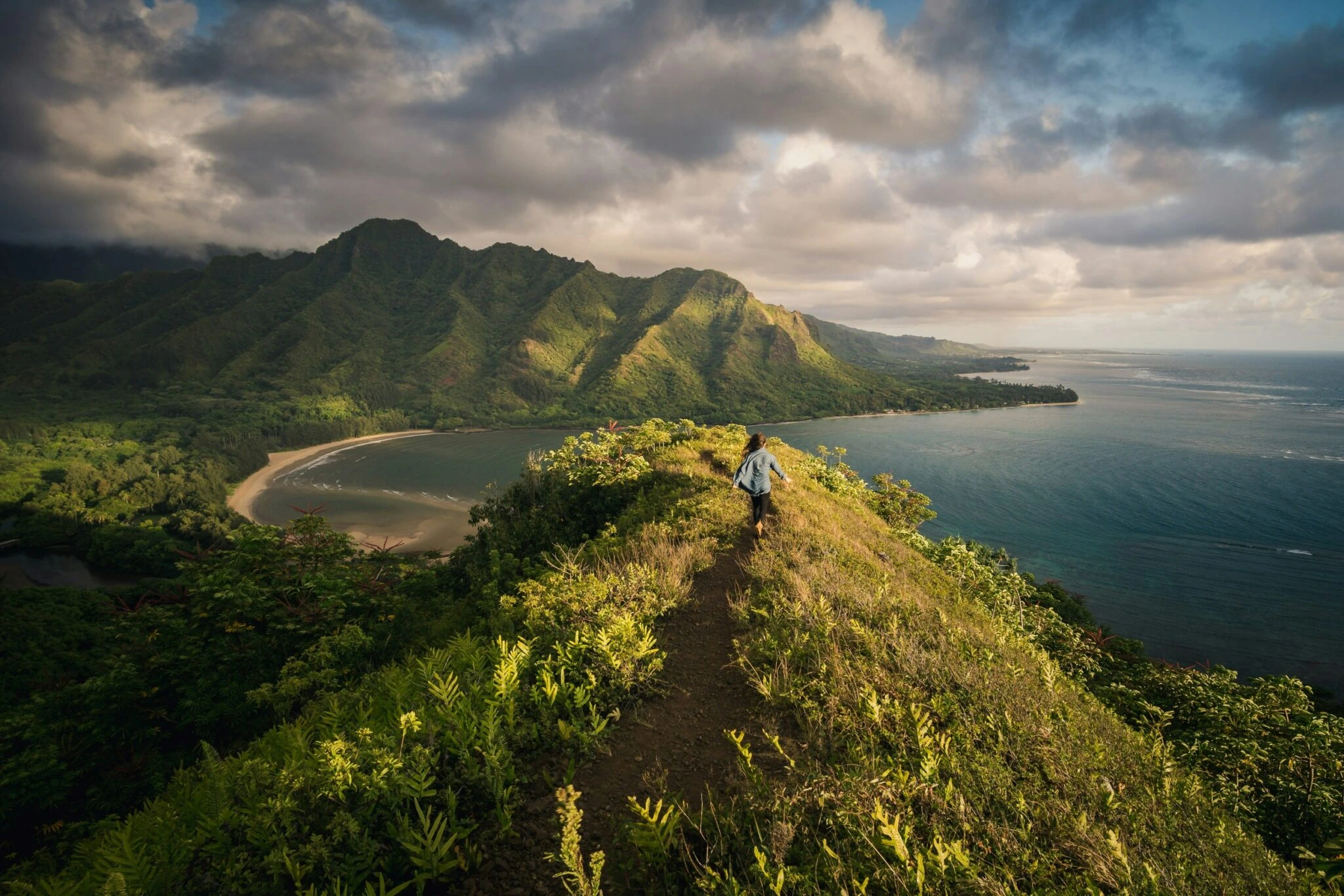 Hiker walking along a lush mountain ridge trail overlooking a tropical bay and dramatic coastal mountain range
