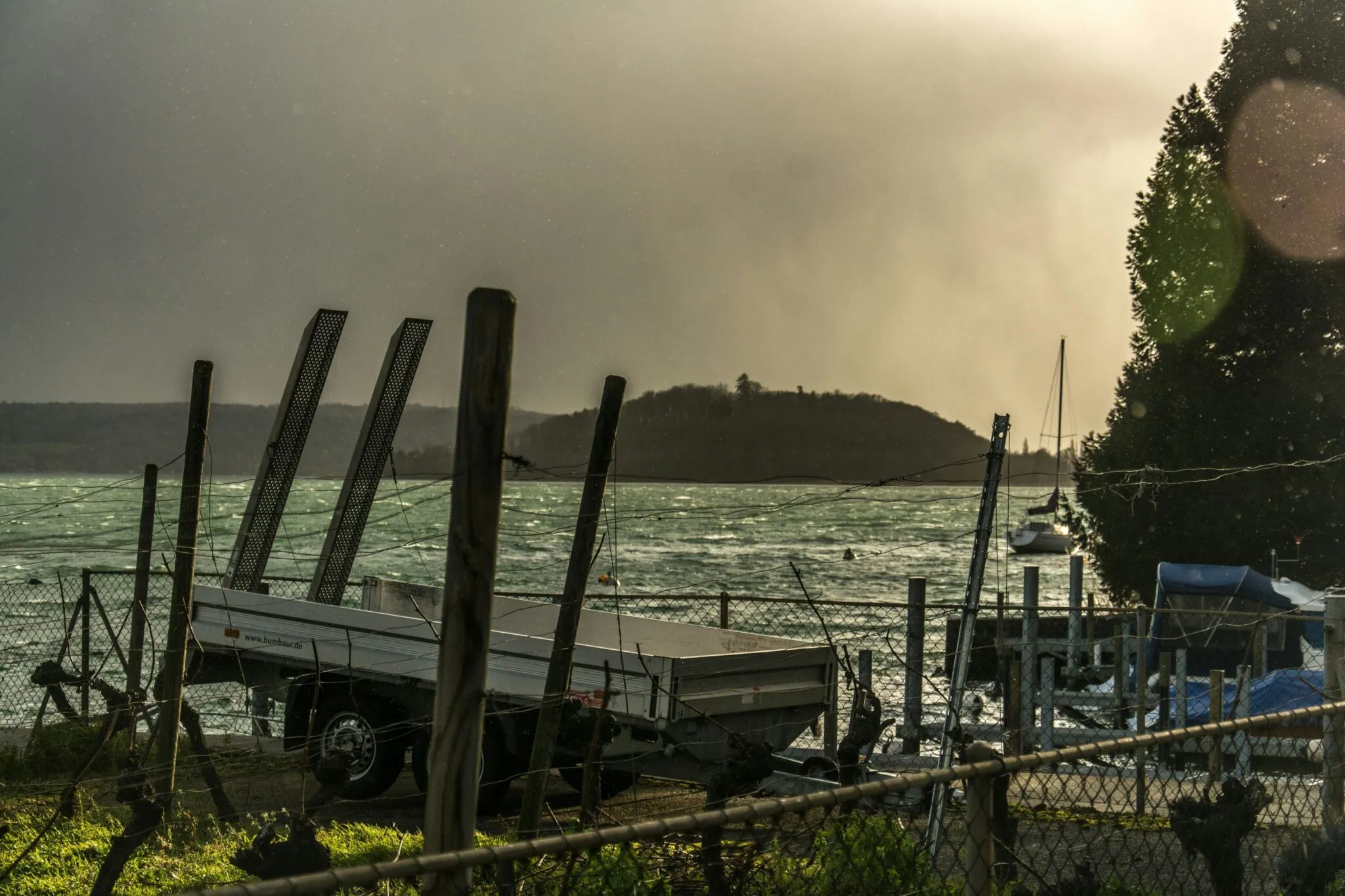 Utility trailer with loading ramps parked behind a fence overlooking a stormy lake with choppy waves and distant sailboat