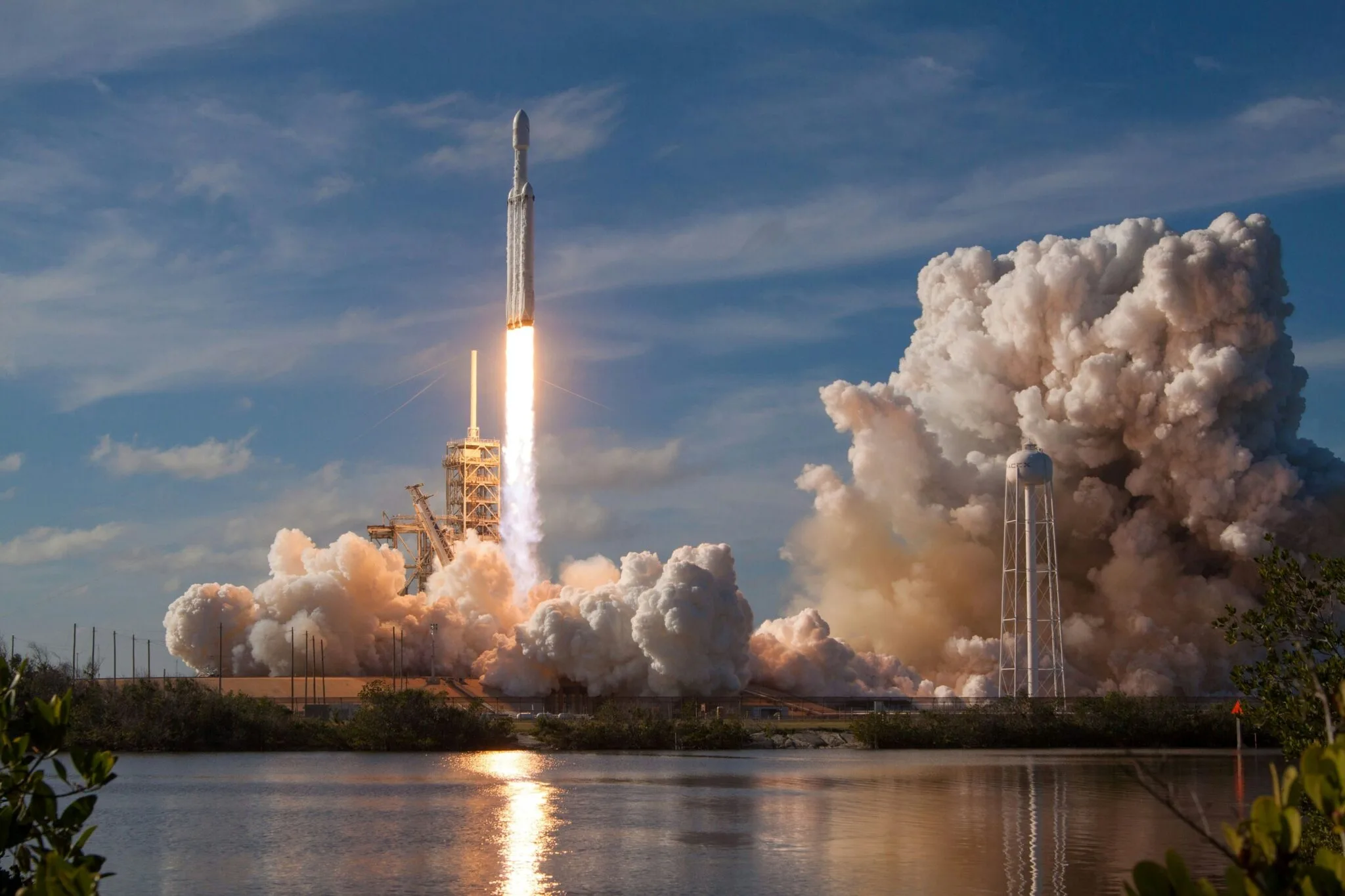 SpaceX Falcon Heavy rocket lifting off from launch pad with massive smoke plume and fiery exhaust reflection over water