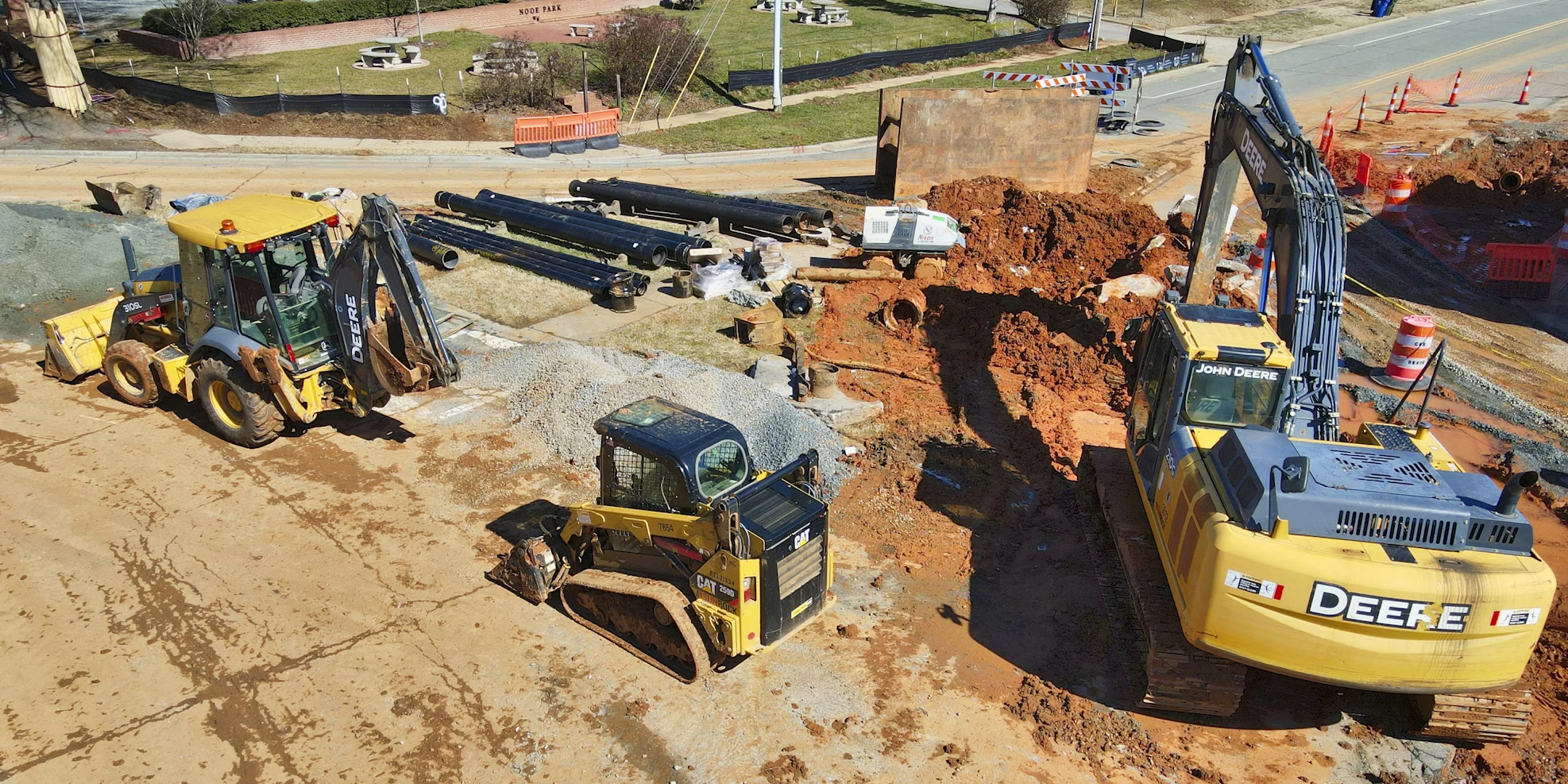 Heavy construction equipment including John Deere excavator, backhoe, and Cat skid steer at a job site