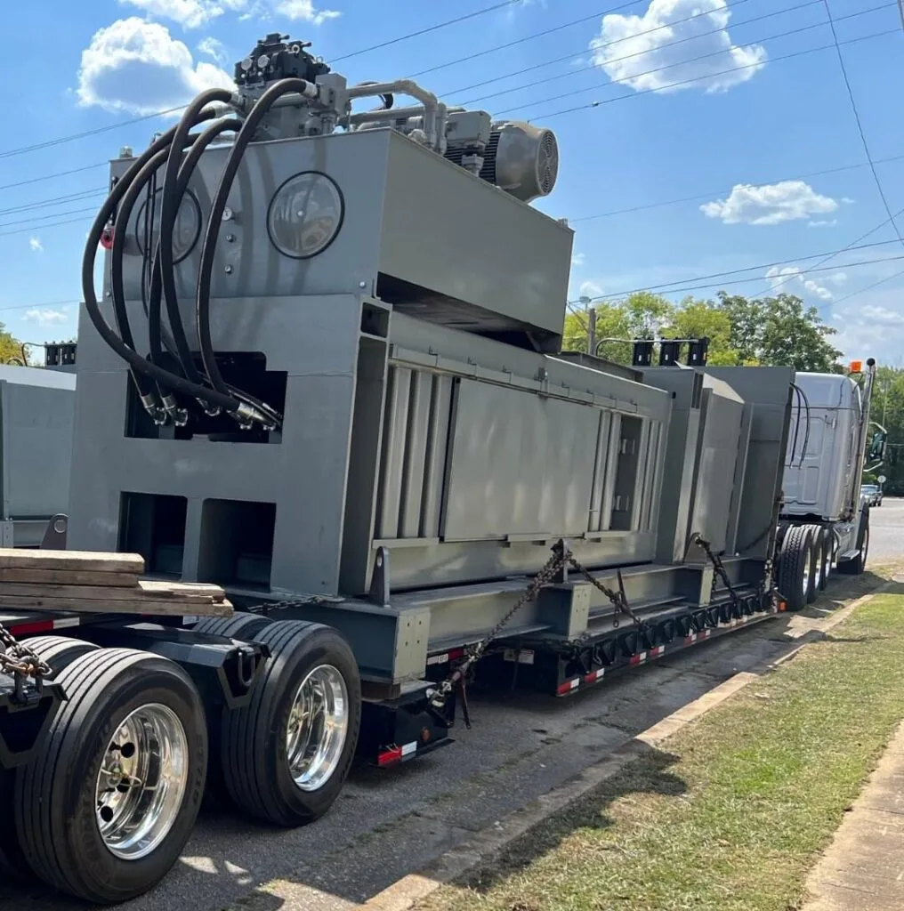 Massive grey industrial hydraulic press machinery chained securely to an RGN lowboy trailer for heavy haul freight shipping