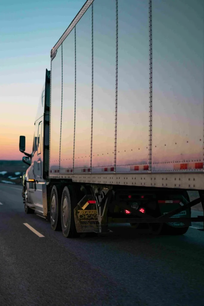 Rear perspective of a commercial semi-trailer truck driving along a highway during a vibrant sunset