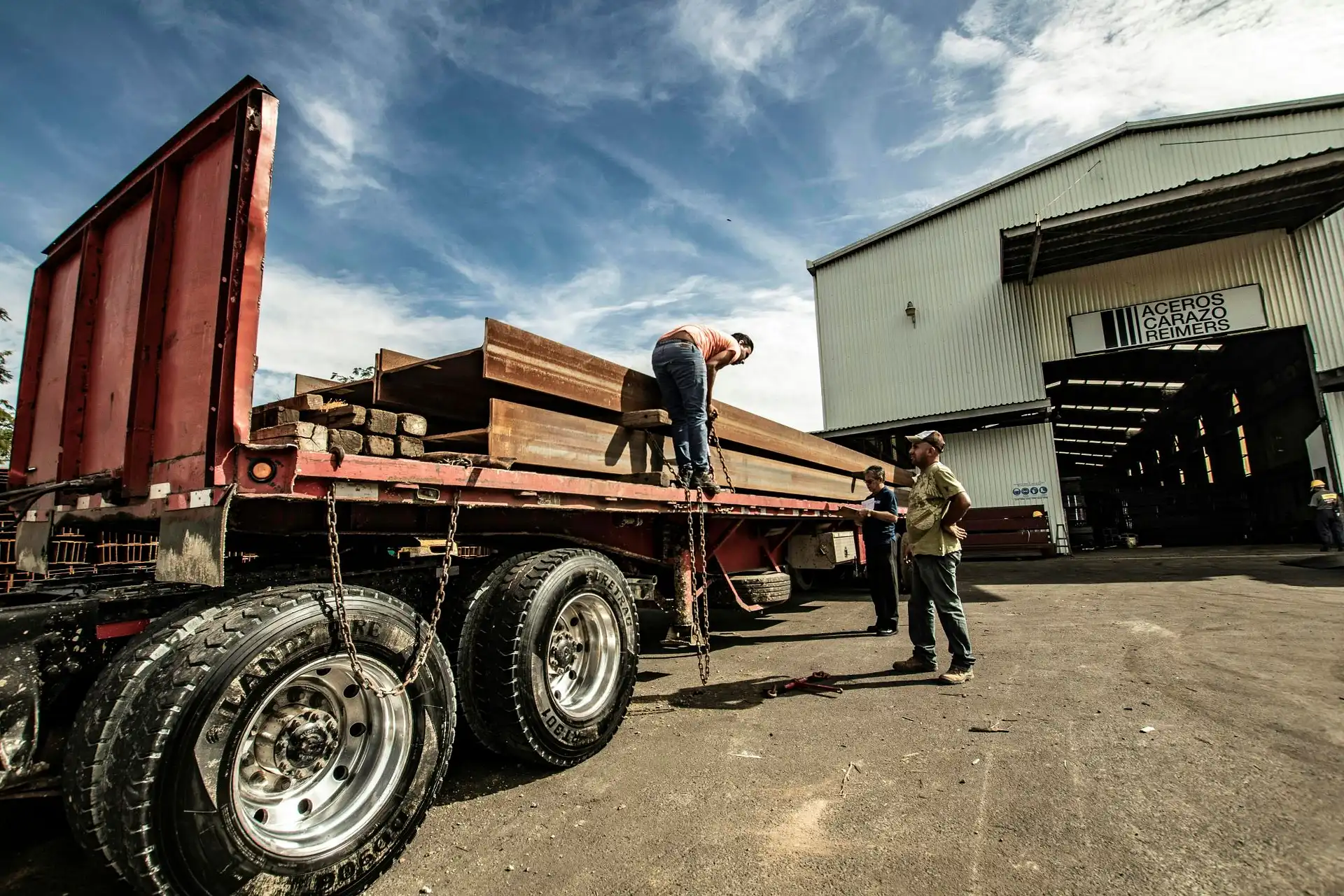 Workers securing heavy structural steel I-beams with transport chains onto a flatbed trailer at an industrial metal fabrication facility