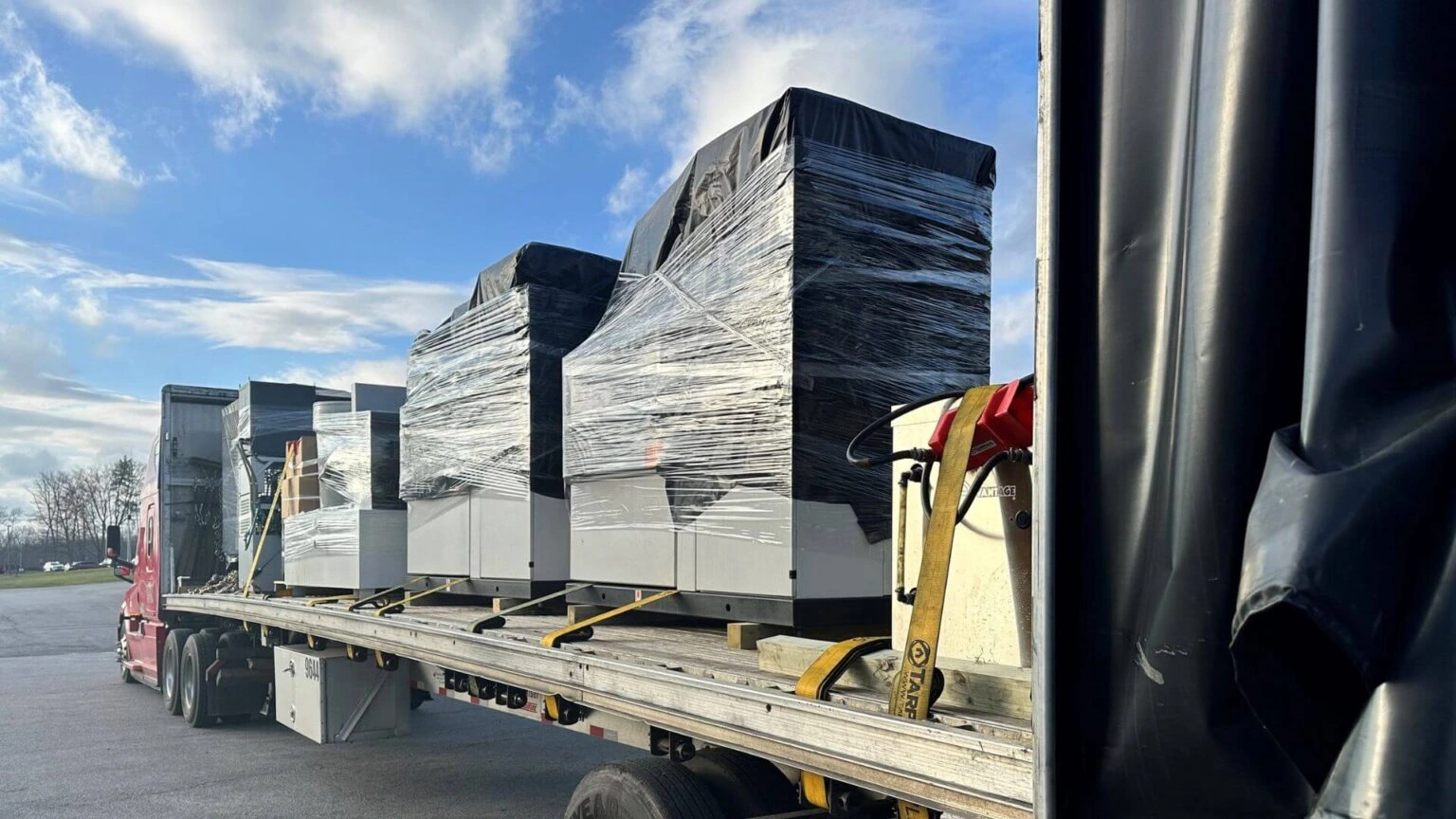Conestoga flatbed trailer hauling shrink-wrapped industrial machinery secured with heavy-duty cargo straps