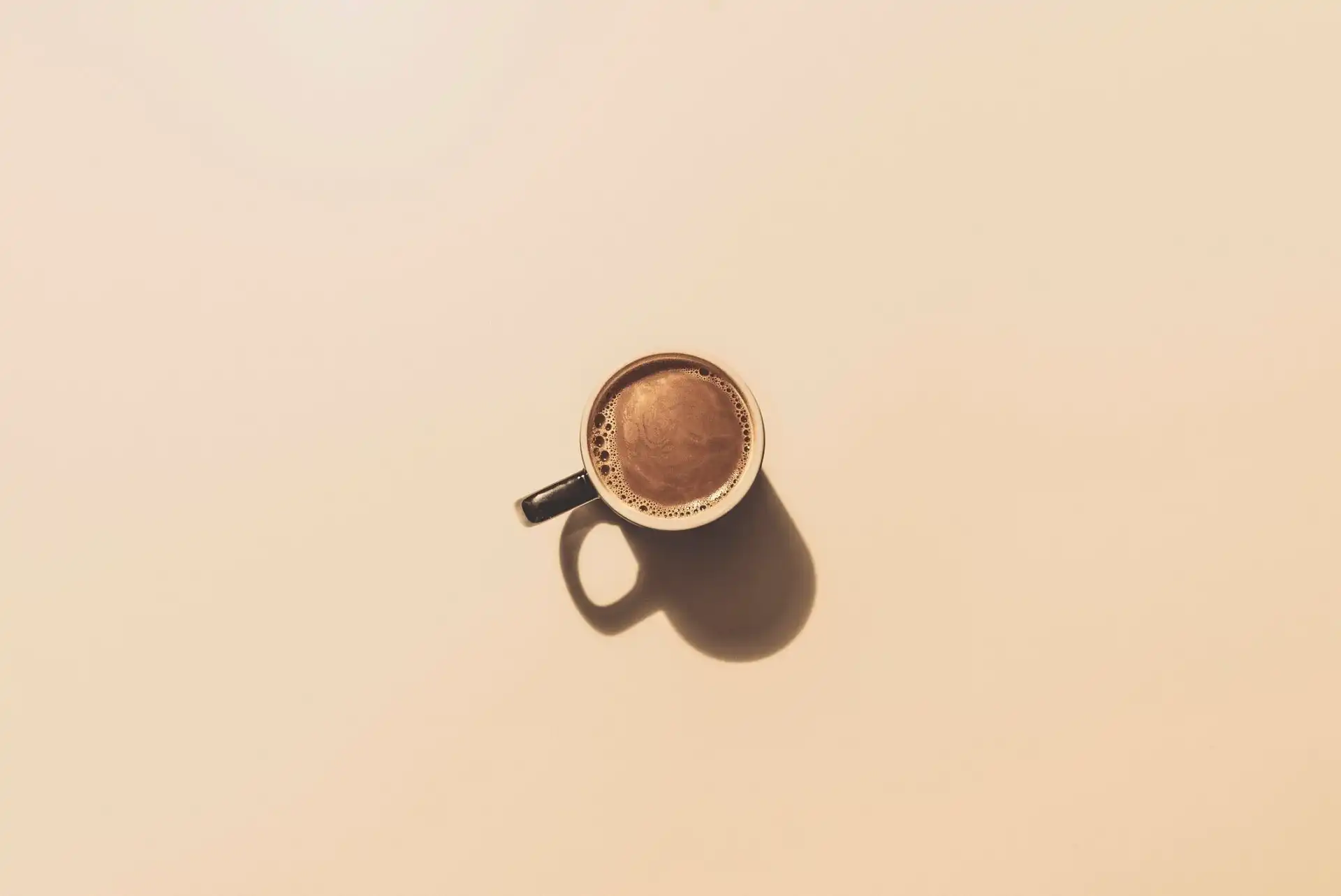 Top-down minimalist view of a fresh cup of coffee casting a soft shadow on a warm neutral desk surface