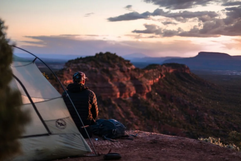 Hiker sitting beside a camping tent on a mountain cliff overlooking a canyon valley at sunset