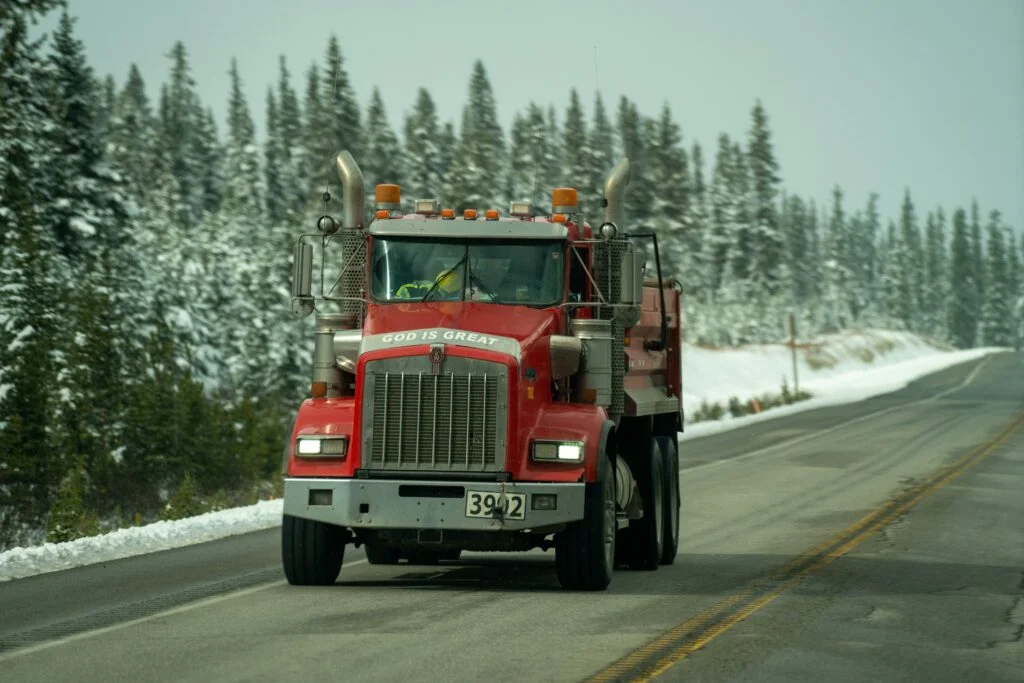 Red Kenworth heavy-duty dump truck driving on a mountain highway with snow-covered pine trees