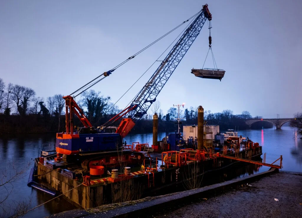 Crawler lattice-boom crane mounted on a floating barge lifting a small workboat from the river