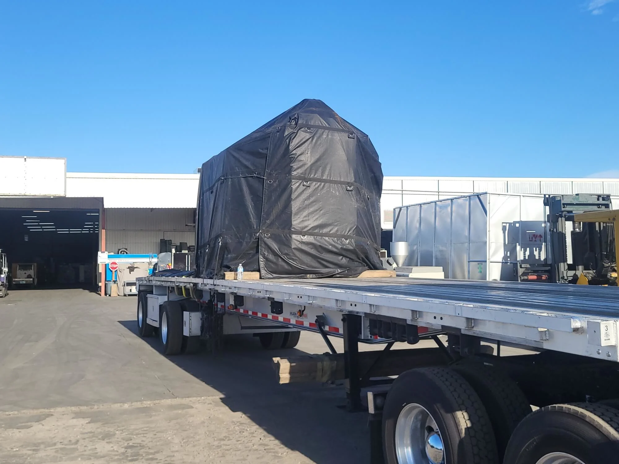 Heavy industrial machinery protected by heavy-duty black weather tarp secured onto an aluminum flatbed semi-trailer